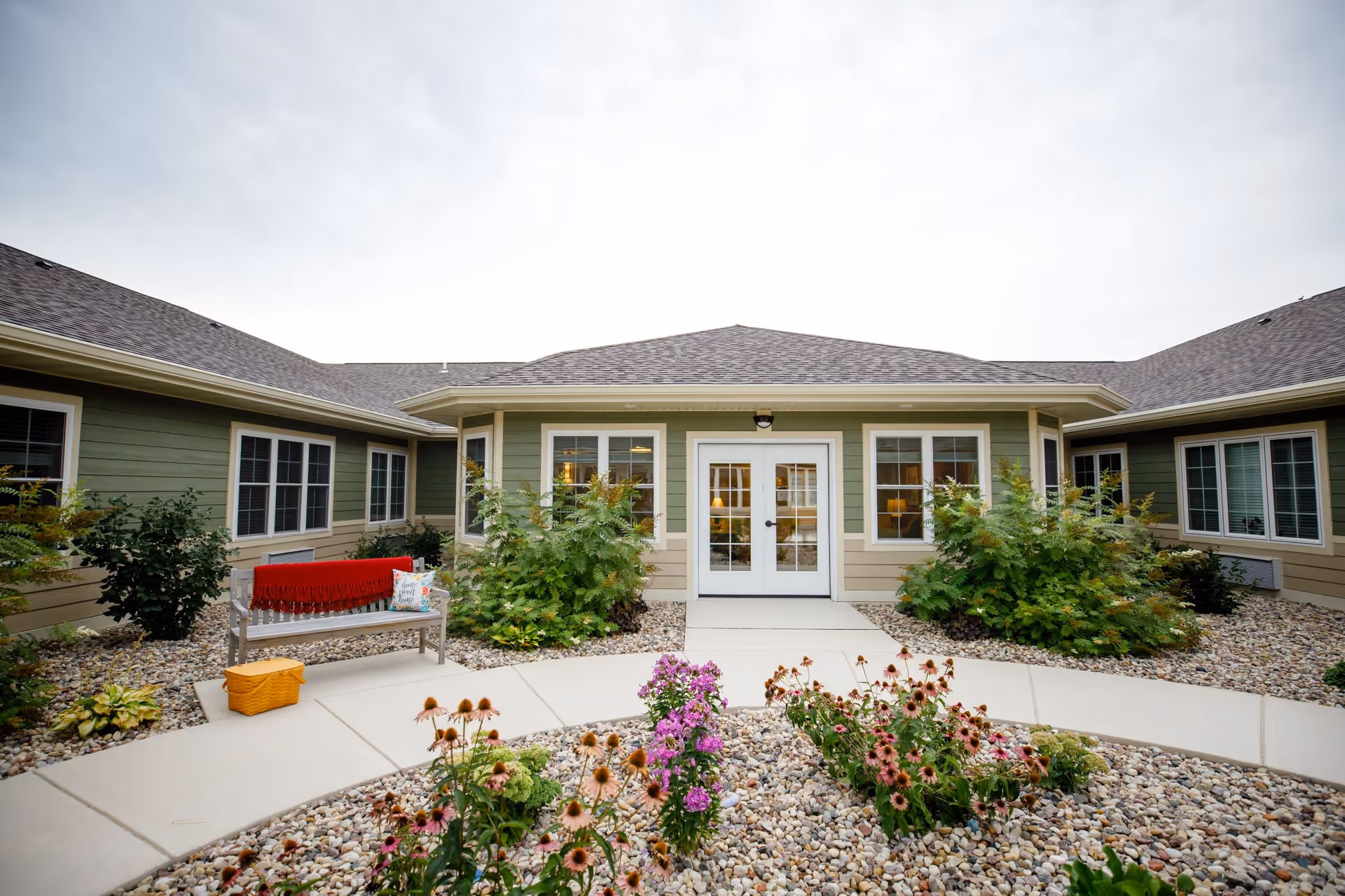 Outdoor courtyard area of The Courtyard at Bellevue Memory Care and Assisted Living, featuring a paved walkway, flower beds with blooming flowers, green shrubs, and a bench with a red blanket and a pillow. The building exterior is light green with beige trim and multiple windows surrounding the courtyard.