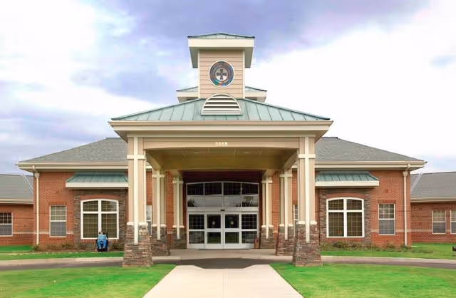 Front exterior view of a single-story brick building with a covered entrance supported by columns. The building has a green metal roof with a small tower featuring a circular window with a cross design. There is a concrete walkway leading to the entrance, and a person in a wheelchair is visible near the left side of the building.