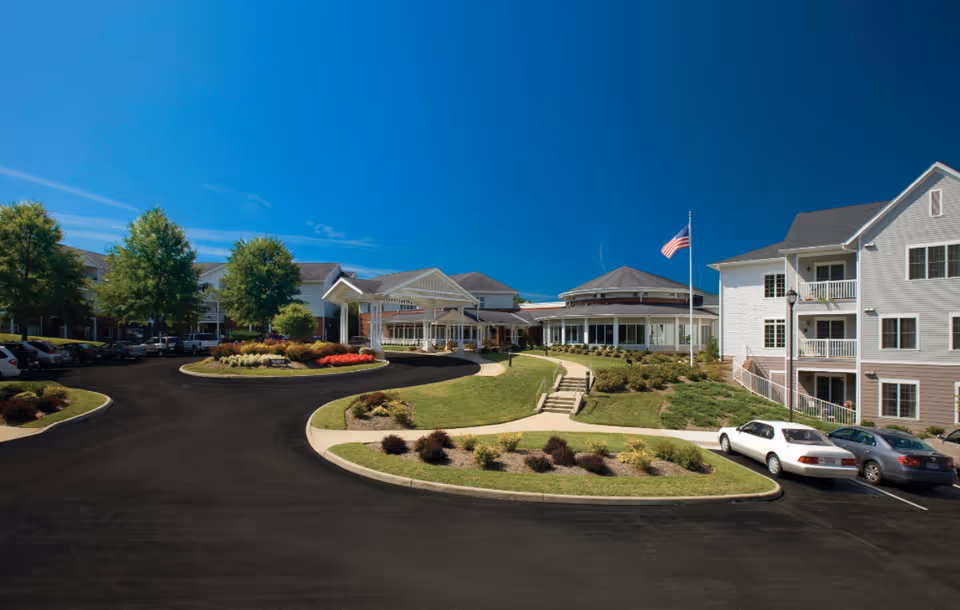 Front entrance and circular driveway of a multi-story nursing and rehabilitation facility with landscaped islands, a flagpole, and parked cars under a clear blue sky.
