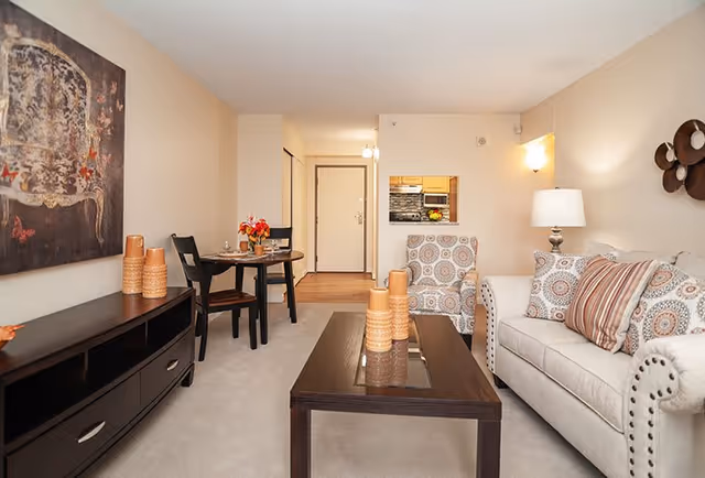 A cozy living room area in a senior living facility featuring a beige sofa with patterned cushions, a matching armchair, a dark wooden coffee table with decorative vases, a small dining table with two chairs, and a console table with additional vases. The walls are light-colored with a large piece of artwork and a decorative wall piece. A lamp is on beside the sofa, and a kitchen pass-through window is visible in the background.