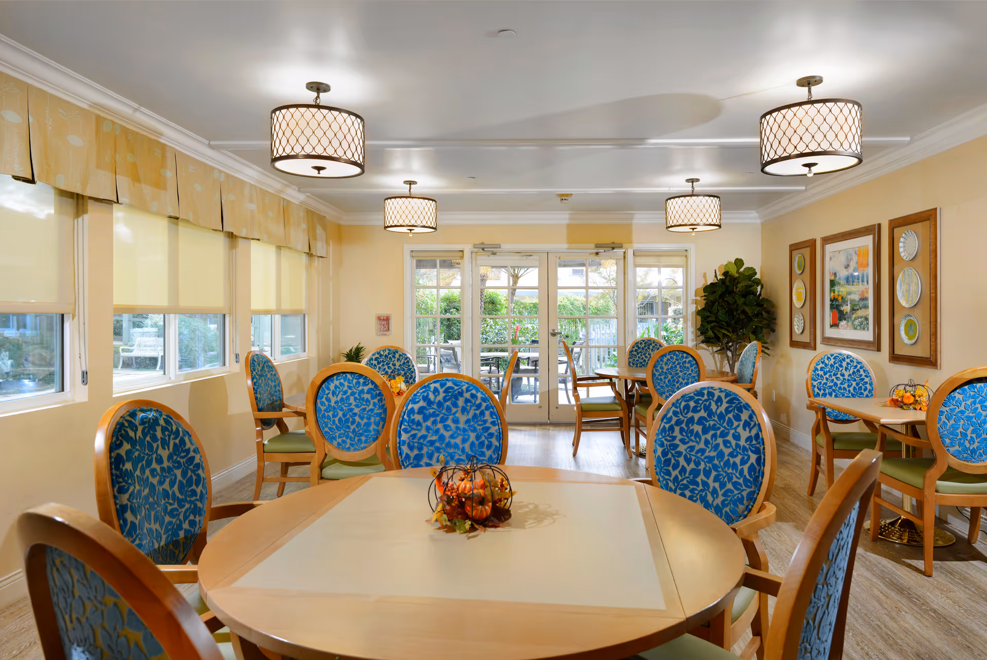 A bright dining room with multiple round wooden tables surrounded by chairs with blue floral upholstery and green cushions. The room has large windows with yellow blinds and valances, three ceiling light fixtures with patterned shades, framed artwork on the walls, and a glass door leading to an outdoor patio area with more seating. There is also a potted plant in the corner.