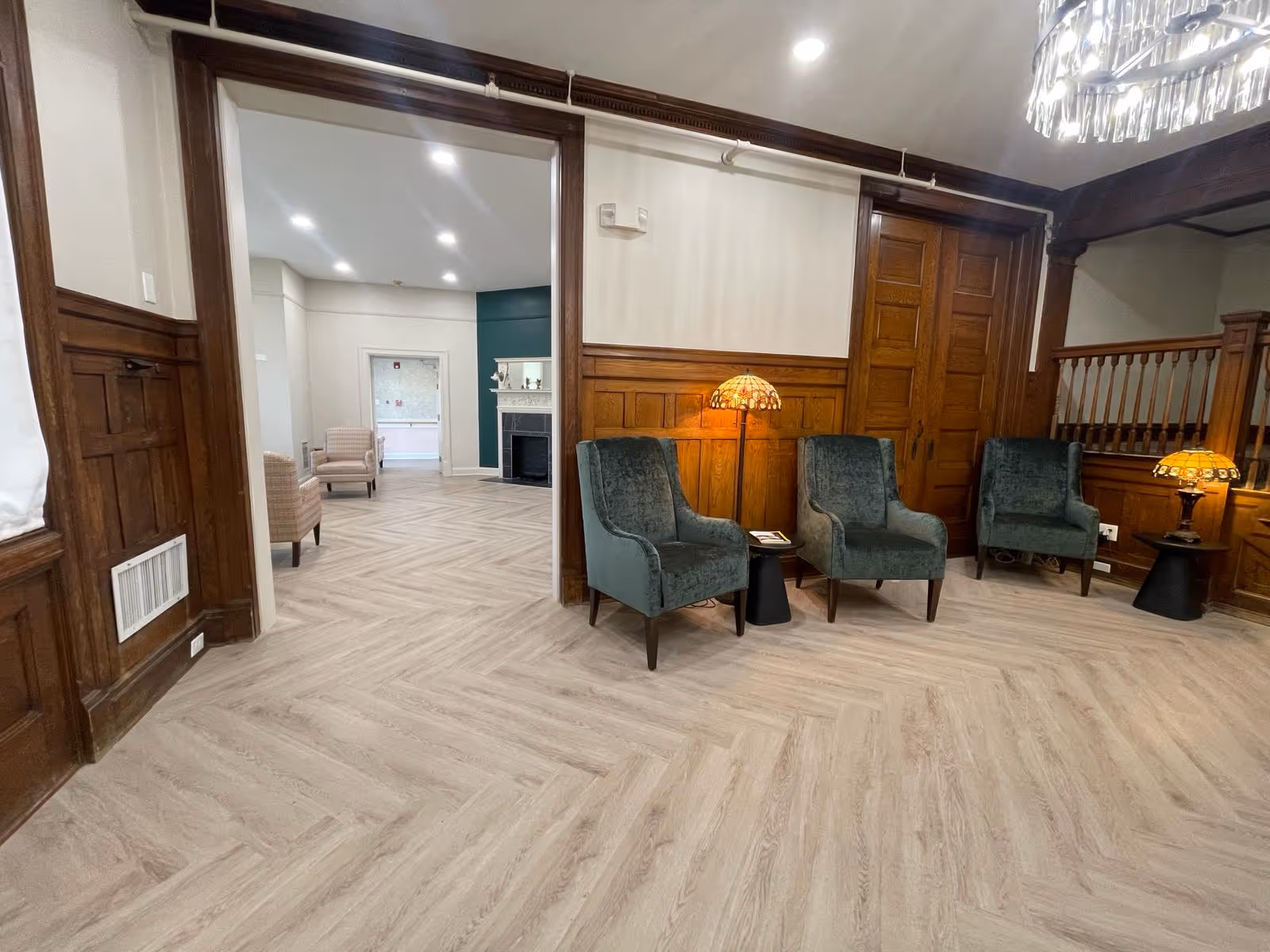 A wood-paneled common room with green upholstered armchairs, Tiffany-style lamps, and light herringbone flooring opening into another seating area.