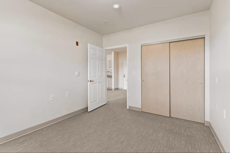 Empty room with beige carpet, white walls, a white door open to an adjacent room, and a wooden sliding closet door.
