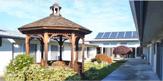 Outdoor courtyard area of Alameda Healthcare & Wellness Center featuring a wooden gazebo surrounded by green bushes and plants, with a concrete walkway and building wings on either side. Solar panels are visible on the roof of the building in the background.