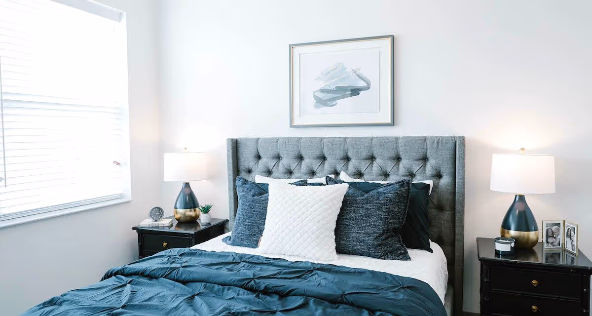 A neatly made bed with a tufted gray headboard, adorned with multiple pillows in white and dark blue. On each side of the bed are black nightstands with matching lamps featuring blue and gold bases. A framed abstract artwork hangs on the white wall above the bed. A window with white blinds is on the left side of the room.