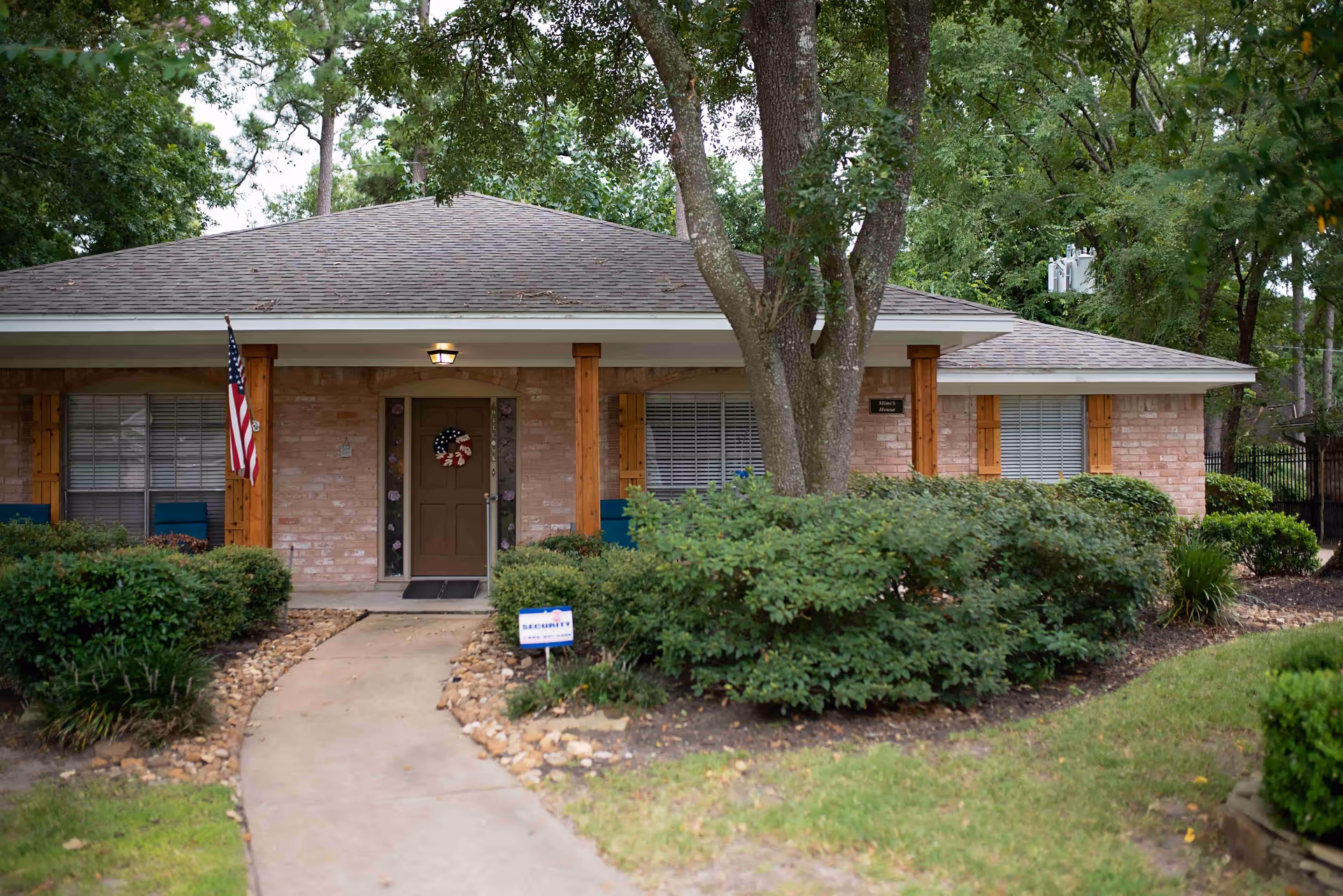 Front exterior of a single-story brick cottage with a walkway, shrubs, an American flag, and a wreath-decorated front door.