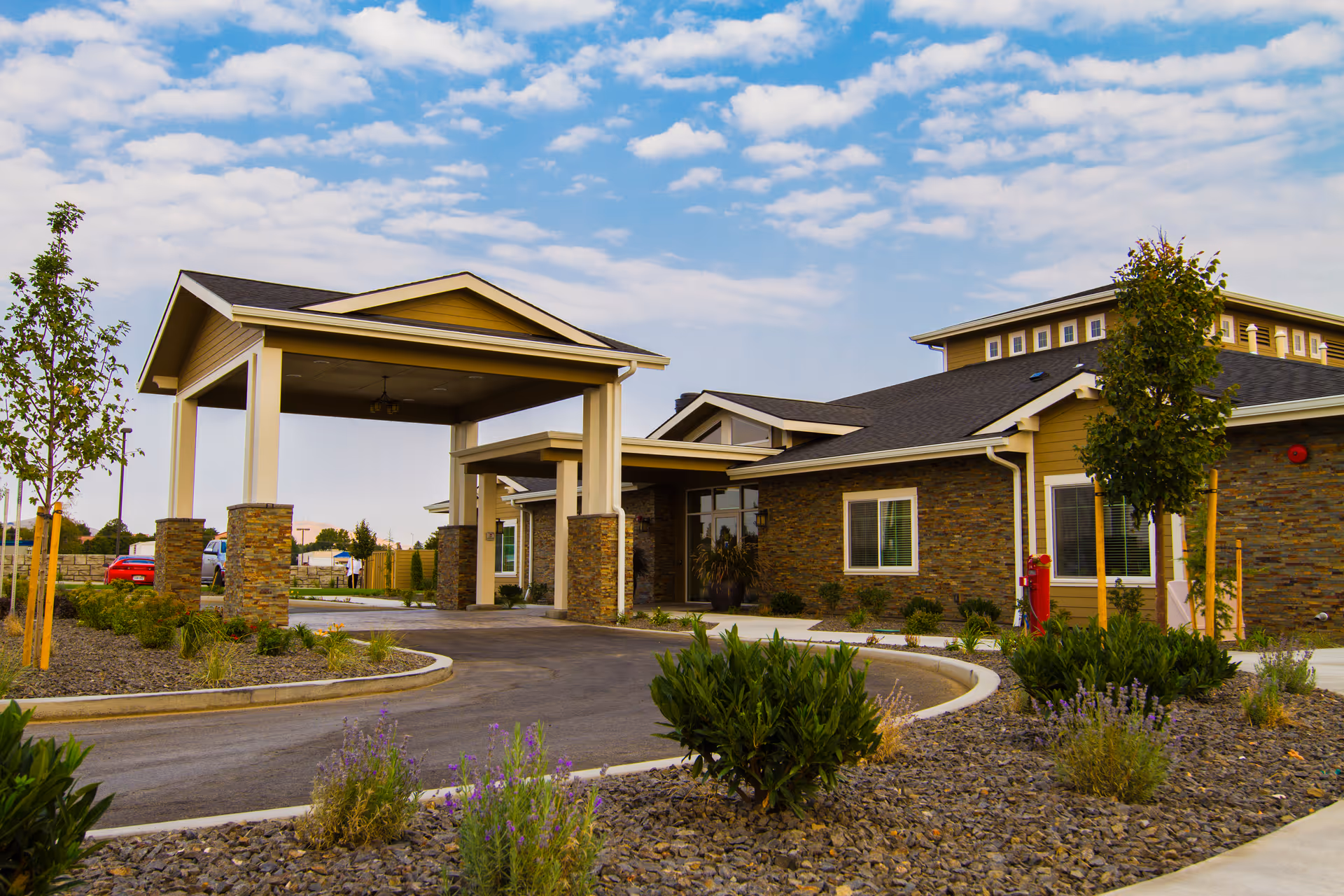Exterior view of a single-story building with a covered entrance supported by stone pillars, surrounded by landscaped plants and trees under a partly cloudy sky.