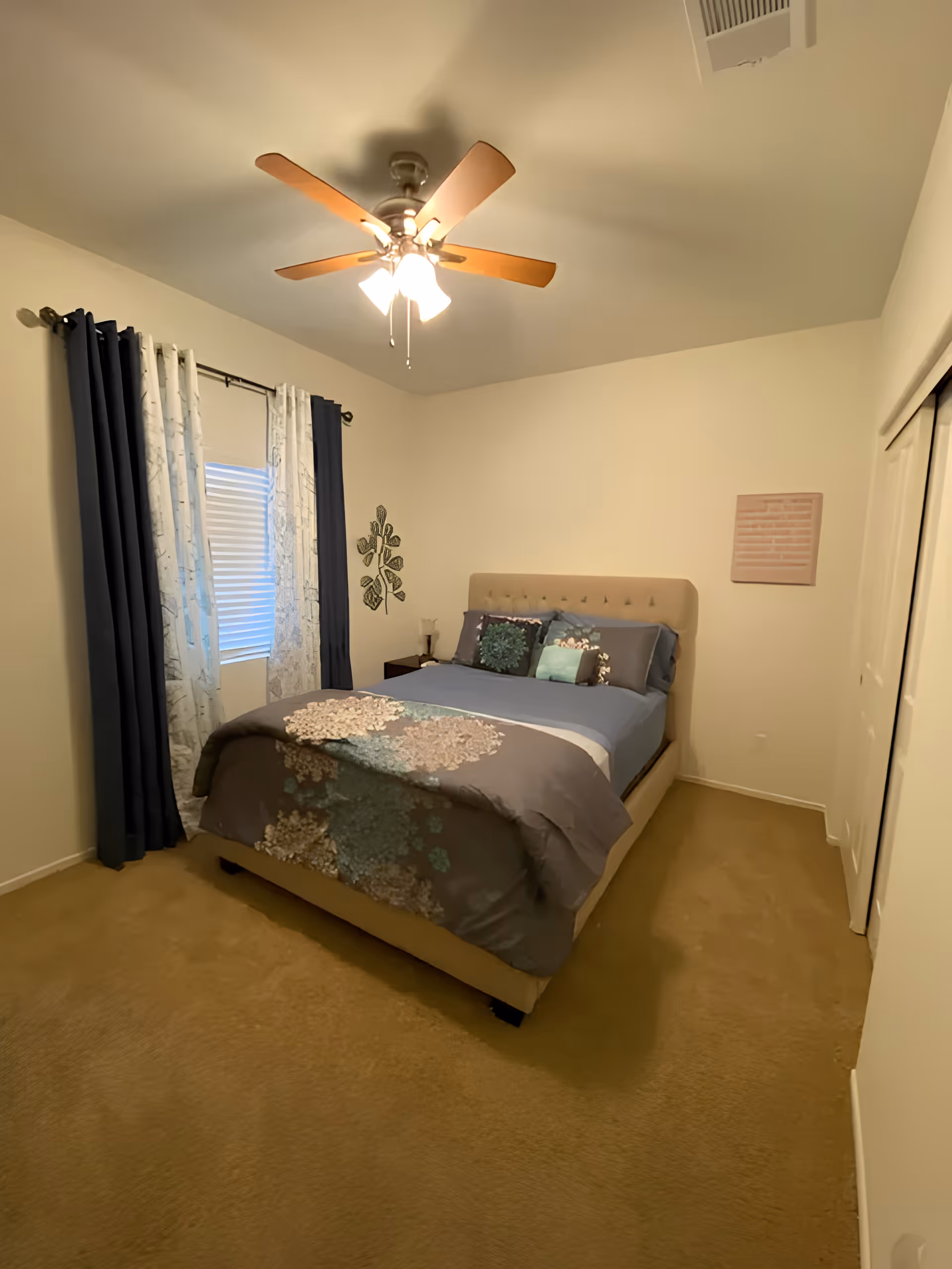 Small bedroom with a tufted bed dressed in patterned bedding, a ceiling fan, curtains over a window, and a nightstand.