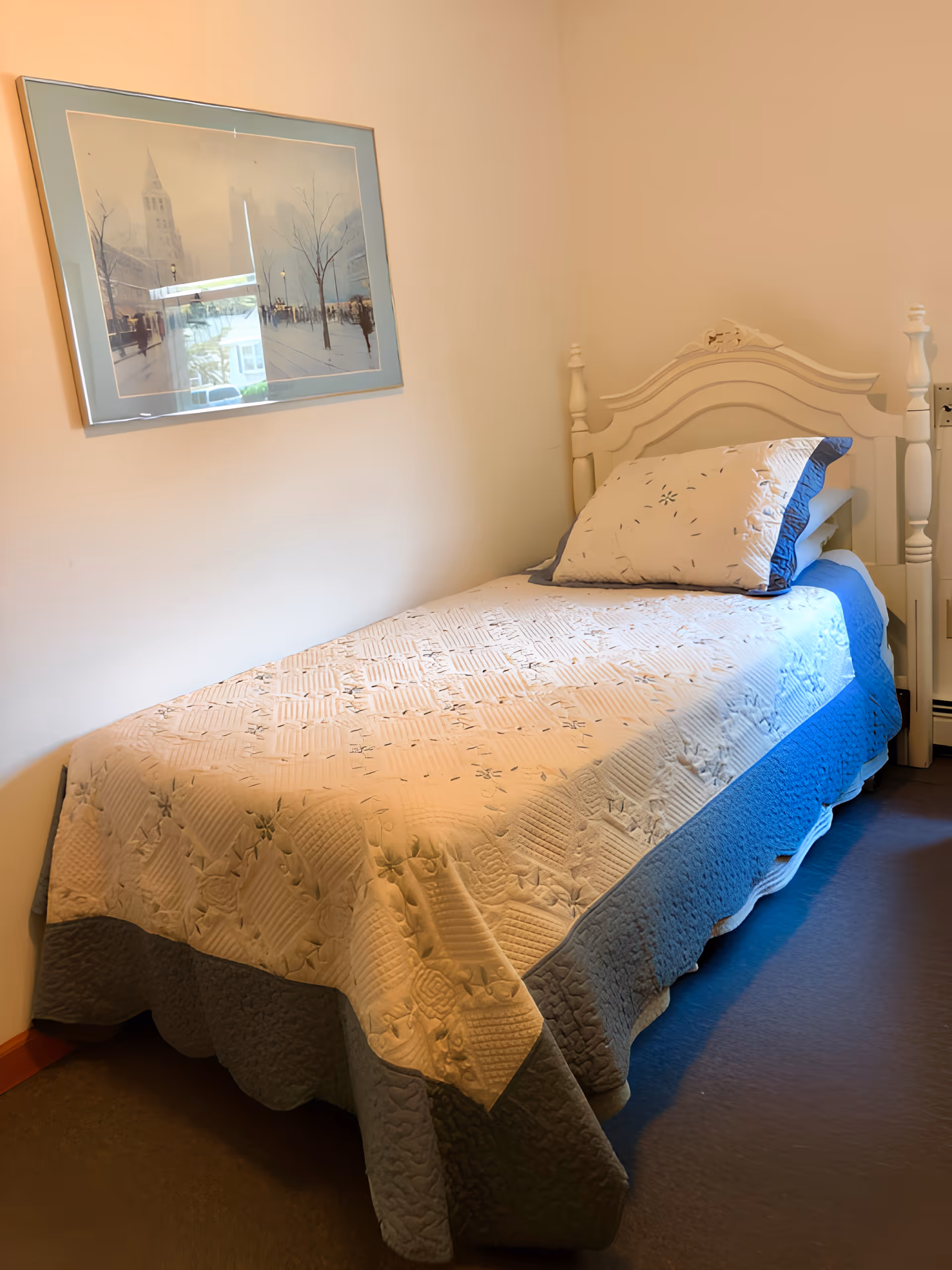 A single bed with a white and blue quilted bedspread and matching pillow in a small room with beige walls. Above the bed, there is a framed picture of a street scene with trees and a tall building.
