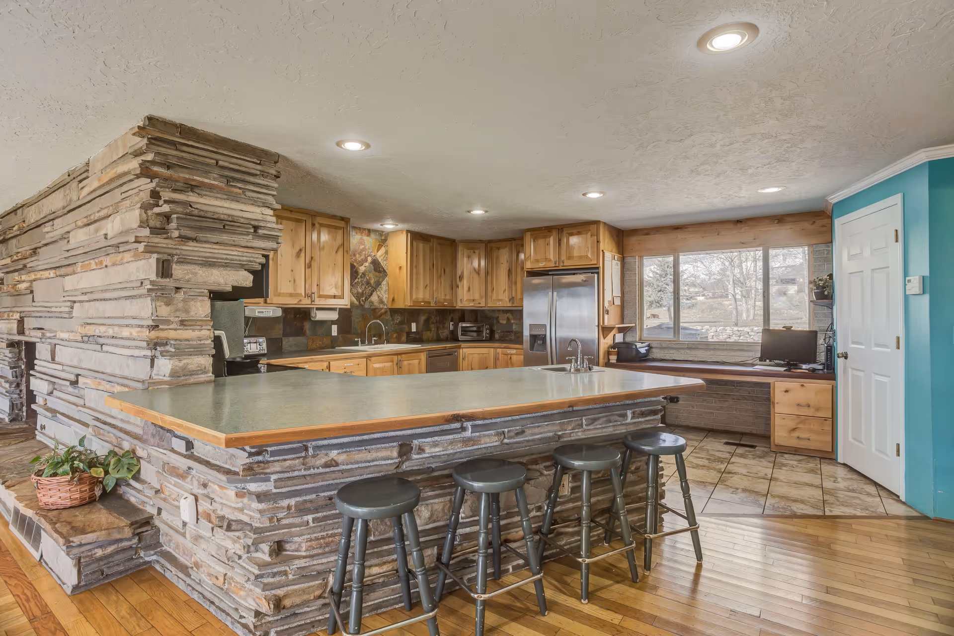 Open kitchen with a stone-faced island and four bar stools, wooden cabinets, stainless refrigerator, and a windowed desk area.