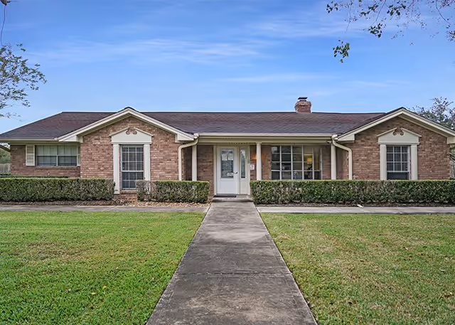 Single-story brick building with a central entrance door, two large windows on either side, a chimney, and a concrete walkway leading up to the entrance. The building is surrounded by neatly trimmed hedges and a well-maintained lawn under a clear blue sky.
