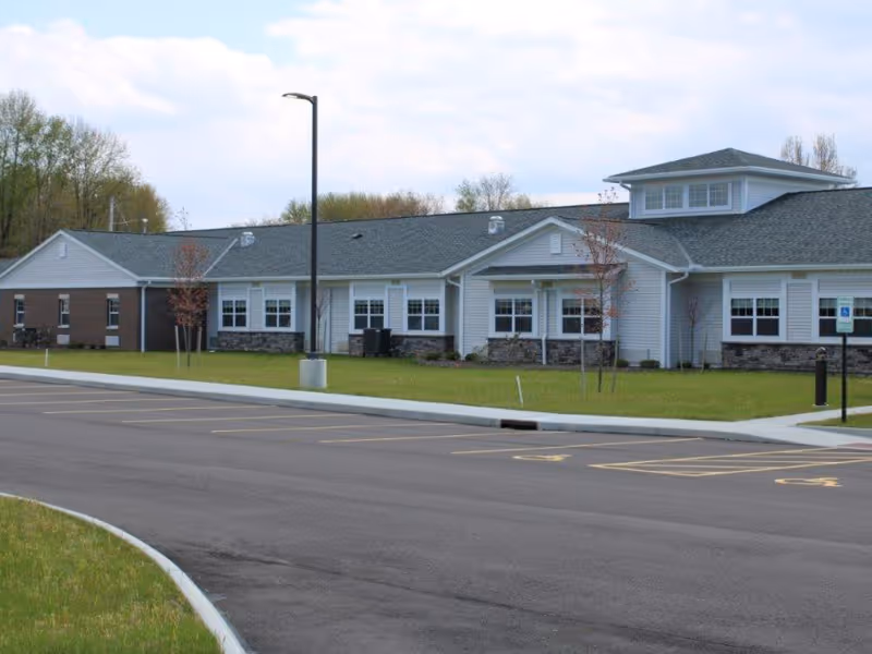 Exterior view of a single-story care and rehabilitation center building with a gray roof, white siding, and stone accents. The building is surrounded by a grassy area with a few small trees and a paved parking lot with marked handicap parking spaces in the foreground.
