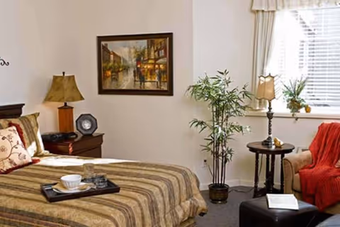 Well-lit bedroom with a made bed, bedside table and lamp, framed painting, potted plant, armchair with a throw, and a window with blinds.