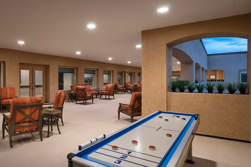 Covered outdoor seating area with multiple cushioned chairs and small tables arranged along the walls. In the foreground, there is a shuffleboard table. The area has beige walls, recessed ceiling lights, and an open window with plants visible outside under a blue sky.