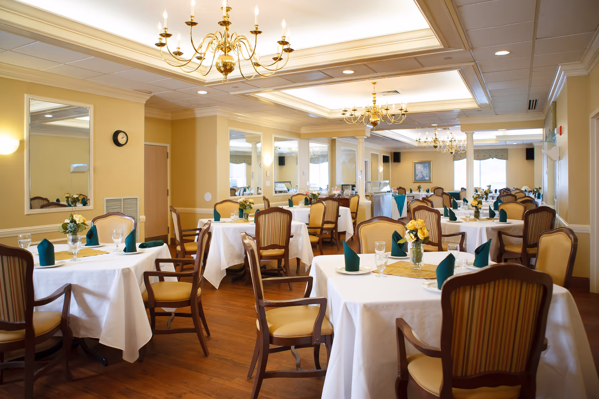 A spacious dining room with multiple tables covered in white tablecloths, each set with green folded napkins, glassware, and small floral centerpieces. The room features wooden chairs with cushioned seats and backs, chandeliers hanging from the ceiling, large mirrors on the walls, and warm yellow paint. The floor is wooden, and the overall ambiance is elegant and inviting.