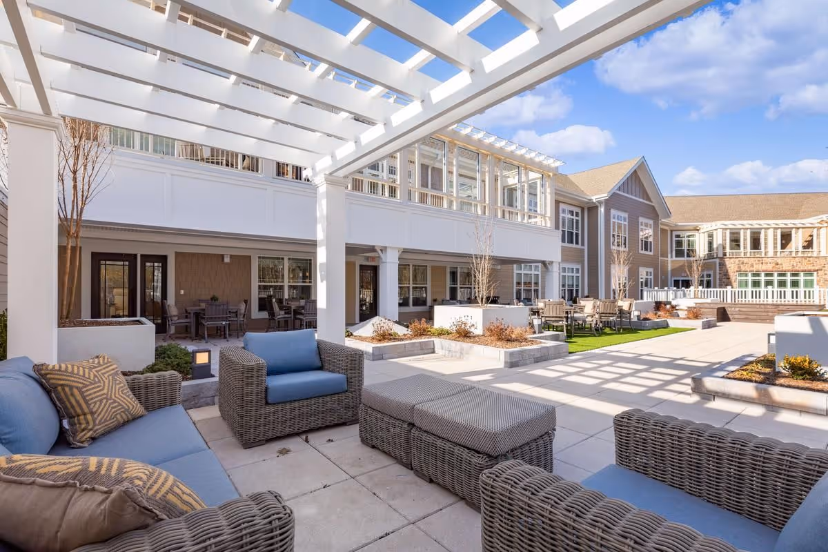 Outdoor patio area at Sunrise of Livingston featuring wicker furniture with blue cushions, a pergola overhead, and a spacious courtyard with tables and chairs surrounded by a multi-story building under a partly cloudy sky.