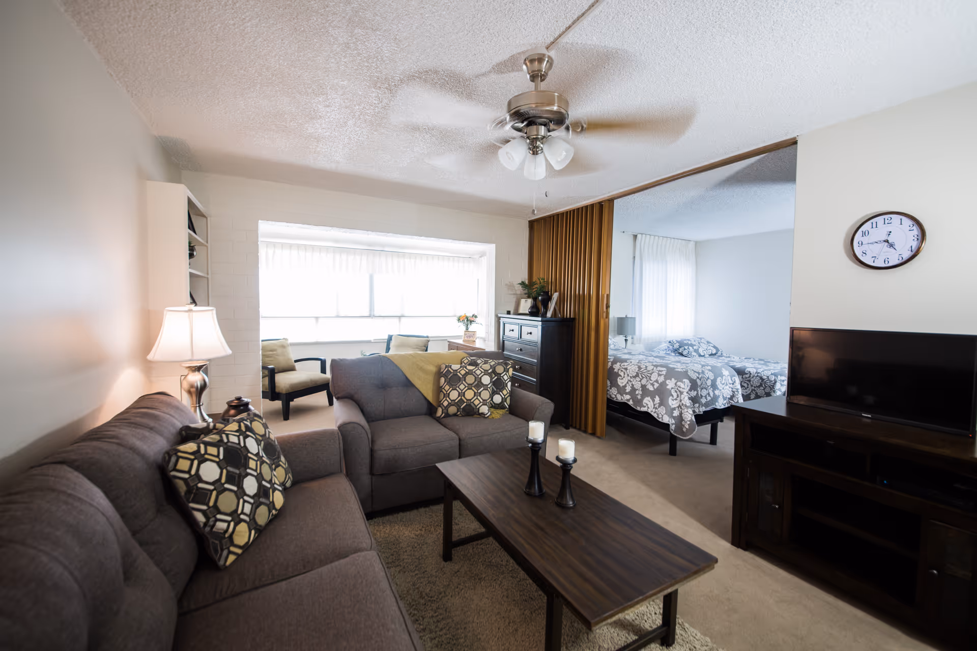 Open living room with sofas, coffee table, TV and an adjacent bedroom visible through a partition.