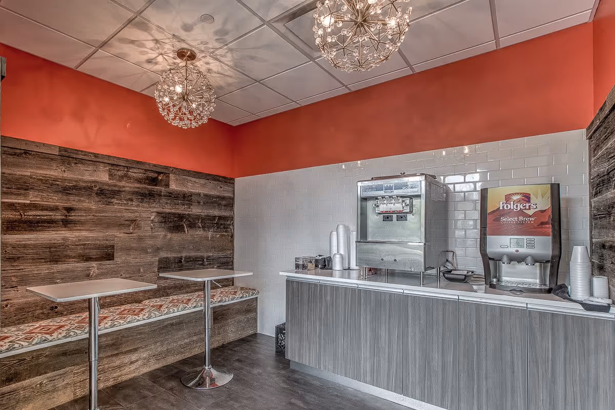 A small dining area with two tables and a bench with patterned cushions along a wooden paneled wall. On the opposite side, there is a counter with a coffee dispenser and a Folgers Select Brew coffee machine. The walls are partly red and white tiled, and two decorative pendant lights hang from the ceiling.