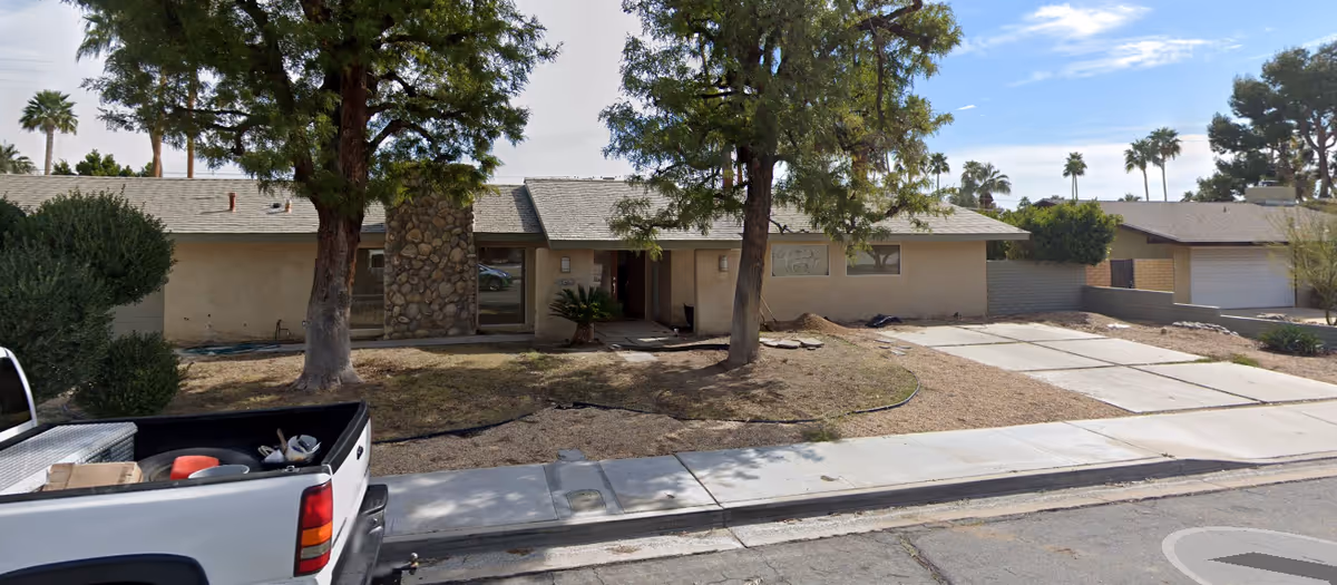 Single-story residential building with a stone chimney, two large trees in front, and a dry lawn area. A white pickup truck is parked on the street in front of the house. The driveway is empty, and there are palm trees visible in the background under a partly cloudy sky.
