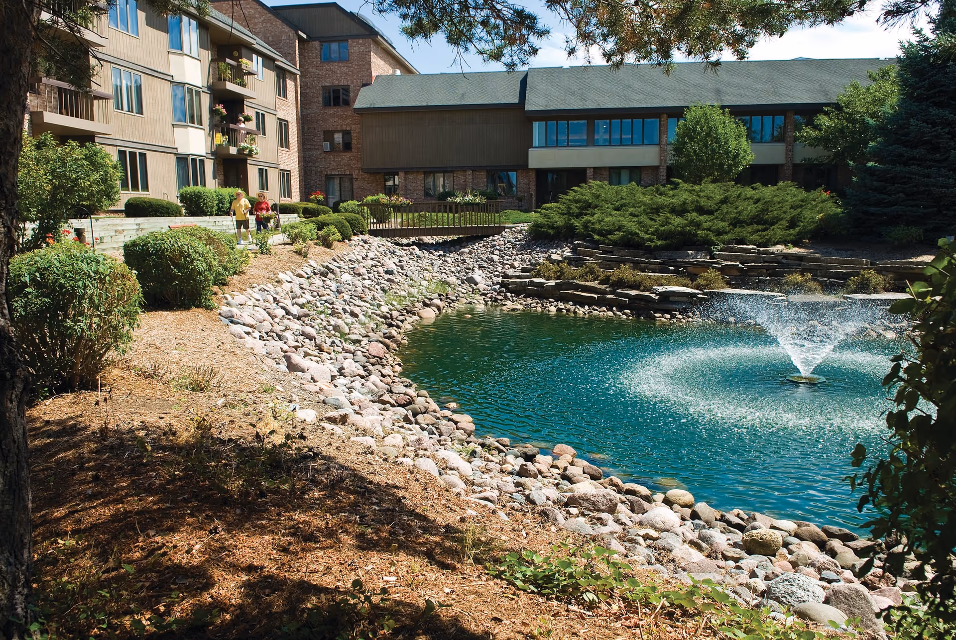 Outdoor view of a senior living facility with a pond featuring a water fountain surrounded by rocks and landscaping. The building has multiple floors with balconies and large windows. Two people are walking on a pathway near the pond.
