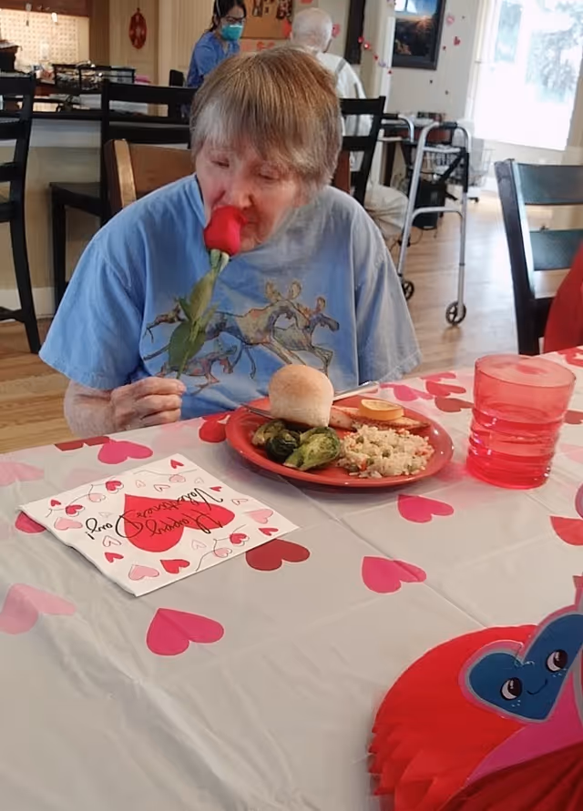 An elderly woman sitting at a table decorated with heart-themed tablecloth and Valentine's Day cards. She is holding and smelling a red rose. On the table in front of her is a red plate with a dinner roll, Brussels sprouts, rice, and a slice of lemon. In the background, a caregiver wearing a mask and another elderly person are visible in a dining area.