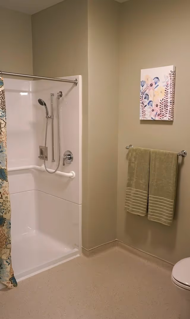 A bathroom with a white shower area featuring a handheld showerhead and a grab bar. There is a floral-patterned shower curtain partially visible on the left. On the right wall, two green towels hang on a towel rack beneath a framed floral artwork. The floor is light-colored and the walls are painted a soft beige.