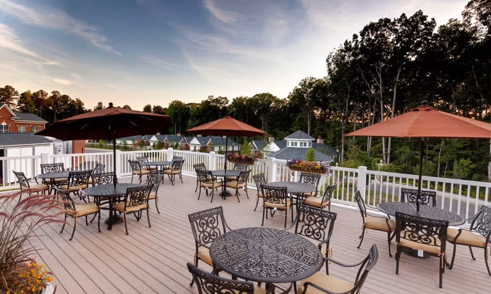 Outdoor patio area with multiple round metal tables and chairs, each table shaded by large brown umbrellas. The patio is surrounded by a white railing and overlooks a wooded area and nearby buildings under a partly cloudy sky at dusk.
