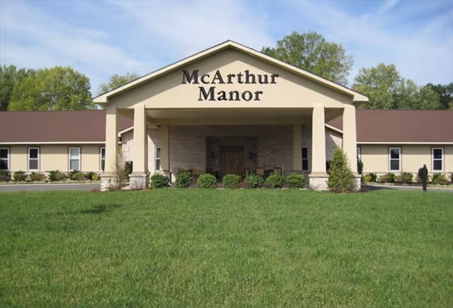 Front exterior view of McArthur Manor Assisted Living building with a large green lawn in front, beige walls, brown roof, and a covered entrance supported by columns.