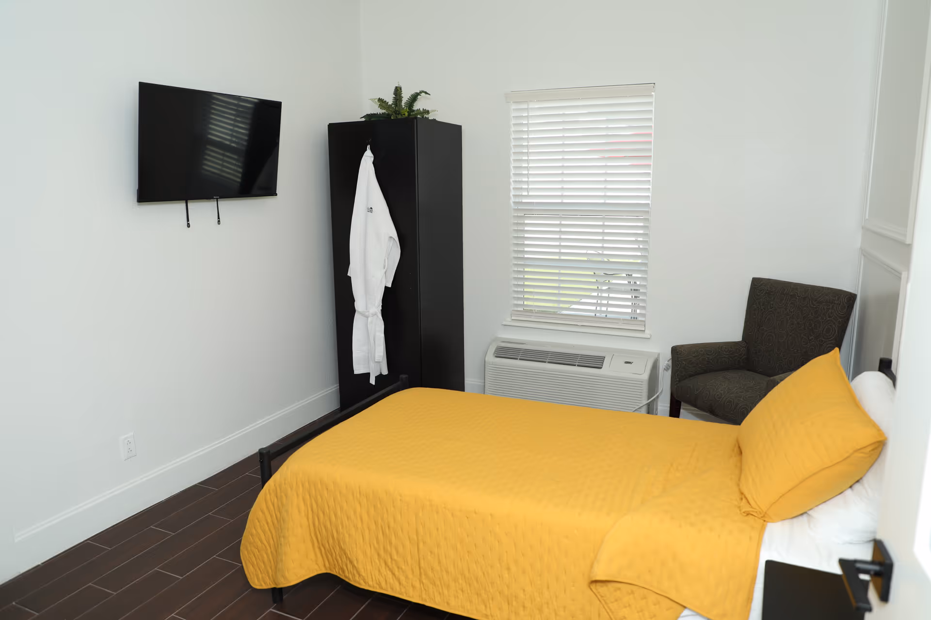 A small bedroom with a single bed covered in a yellow quilt and matching pillow. There is a dark armchair in the corner next to a window with white blinds. A black wardrobe stands against the wall with a white robe hanging on it. A flat-screen TV is mounted on the wall opposite the bed. The floor is dark wood, and the walls are white.