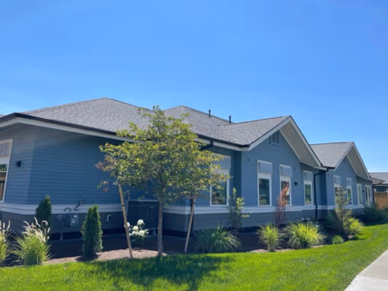 Single-story blue-gray building with landscaped shrubs and a green lawn under a clear blue sky.