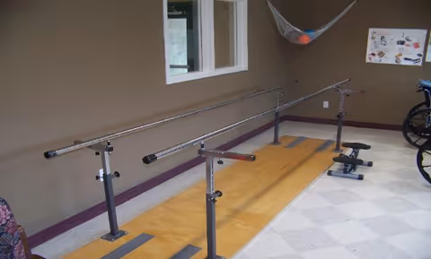 Physical therapy room with parallel bars mounted on a wooden platform for walking exercises, a small stepper machine, a wheelchair, and a netted basket with balls hanging on the wall. A window and a poster with exercise illustrations are also visible.