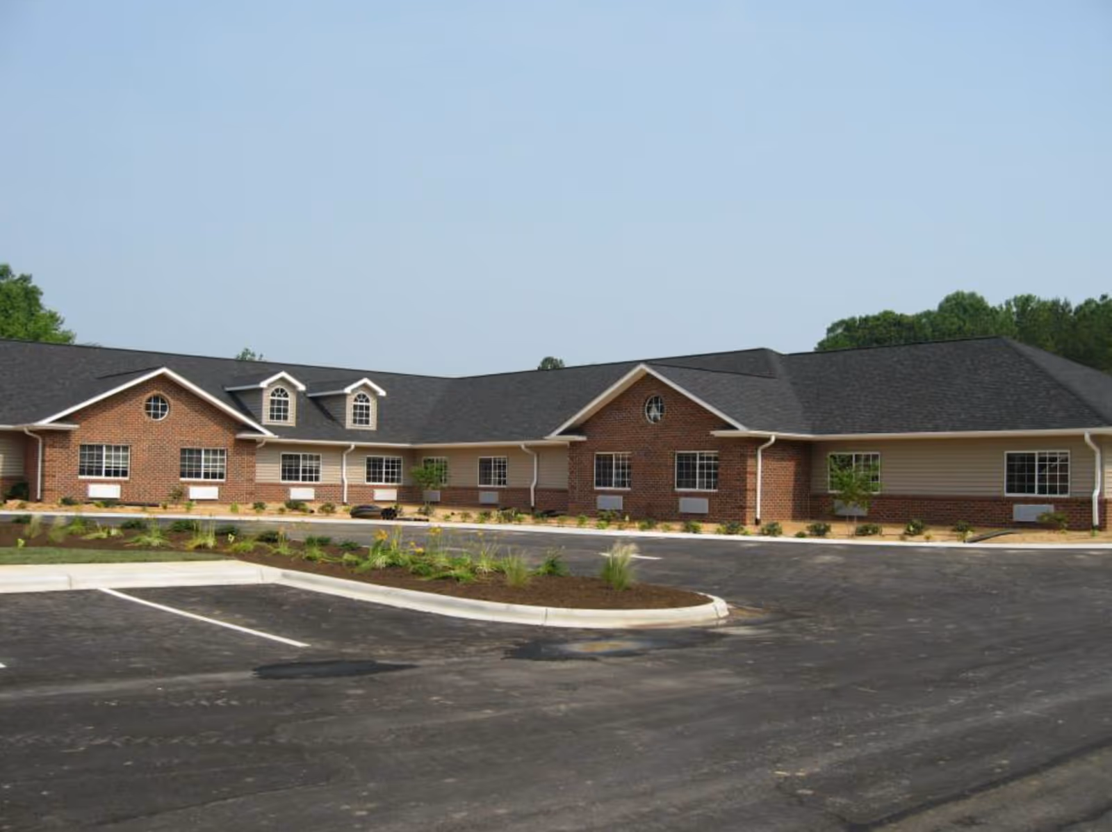 Exterior view of a single-story brick and siding building with multiple windows and a black shingled roof, surrounded by a newly paved parking lot and landscaped areas with small plants and shrubs under a clear sky.