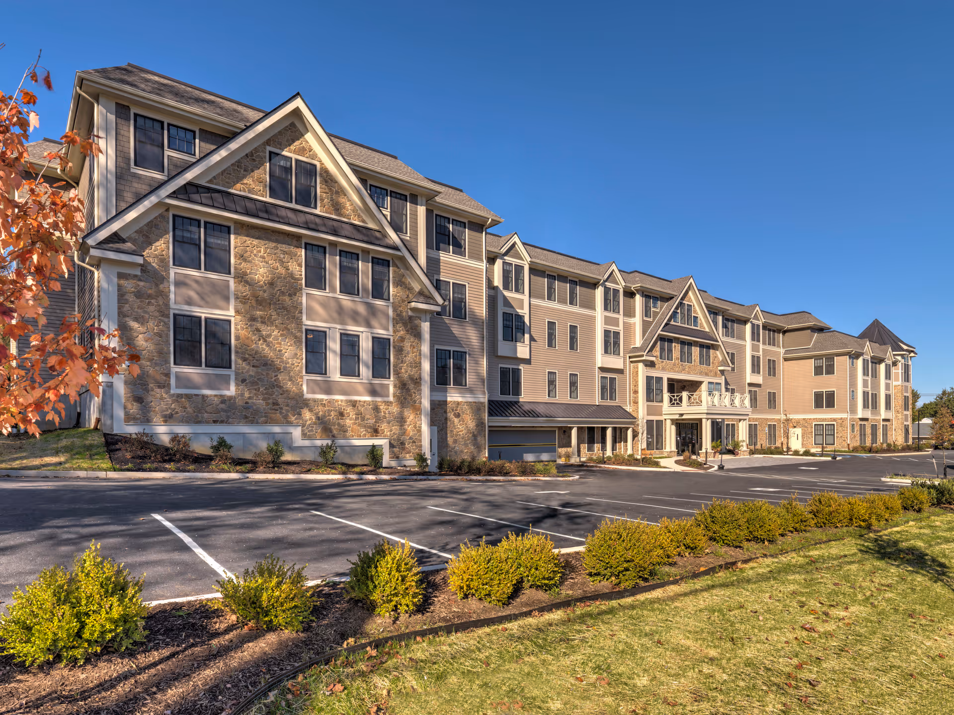 Exterior view of a large, multi-story senior living facility building with stone and siding facade, multiple windows, and a parking lot in front. The sky is clear and blue, with some landscaping including bushes and a tree with autumn leaves.