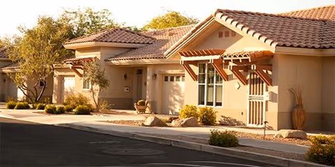 Exterior view of a single-story residential building with beige stucco walls and red tile roofing, surrounded by desert landscaping including small trees and shrubs, under a clear sky.
