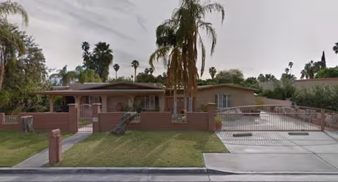 Single-story brick senior care home with a gated front yard and palm trees.
