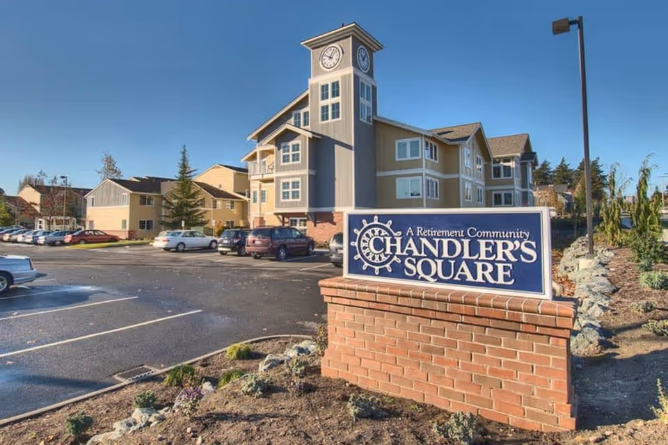 Exterior view of Chandler's Square, a retirement community building with a clock tower. The image shows a parking lot with several cars and landscaped areas with plants and rocks under a clear blue sky.