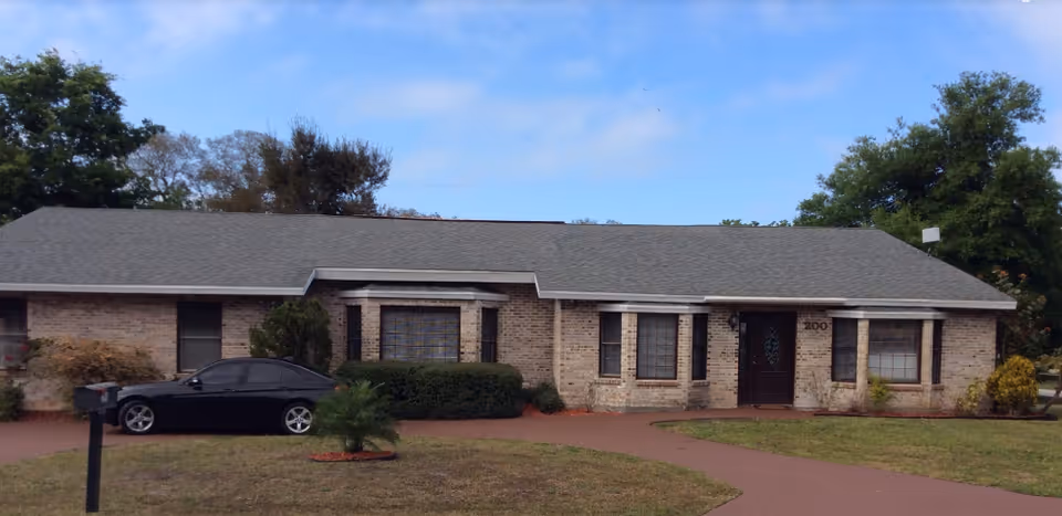 Single-story brick building with a gray shingled roof, several windows, a dark front door, and a driveway with a black car parked. The building is surrounded by grass, small bushes, and trees under a partly cloudy blue sky.