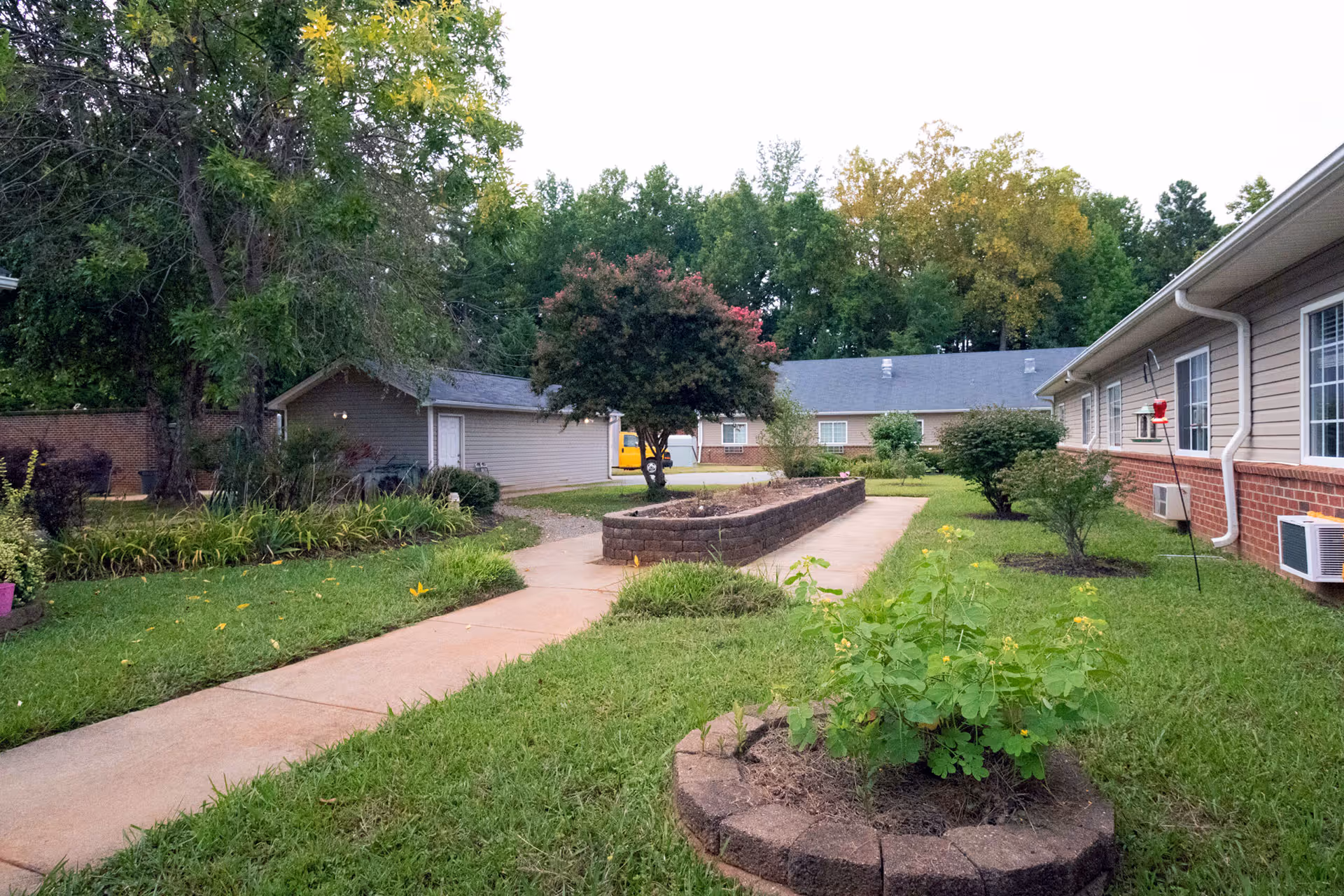 Exterior view of a senior living campus with a central walkway, green lawn, and landscaped garden beds along the building.