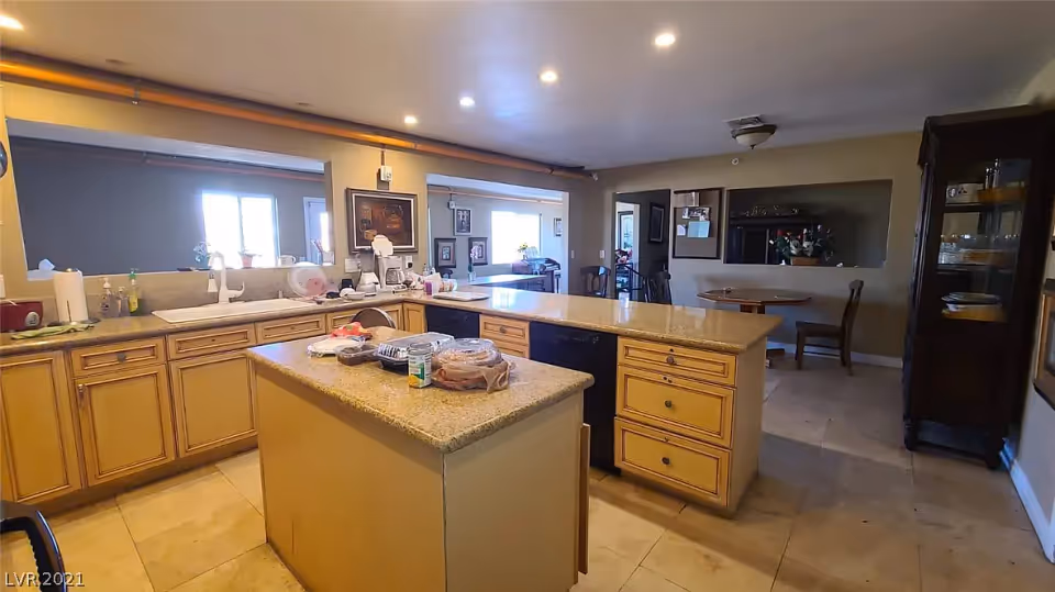 A spacious kitchen area with beige cabinets and granite countertops. There is a central island with various food items on it. The kitchen has a sink, dishwasher, and several kitchen appliances. In the background, there is a small dining area with a round table and chairs, and a glass-front cabinet displaying dishes. The room is well-lit with recessed ceiling lights.