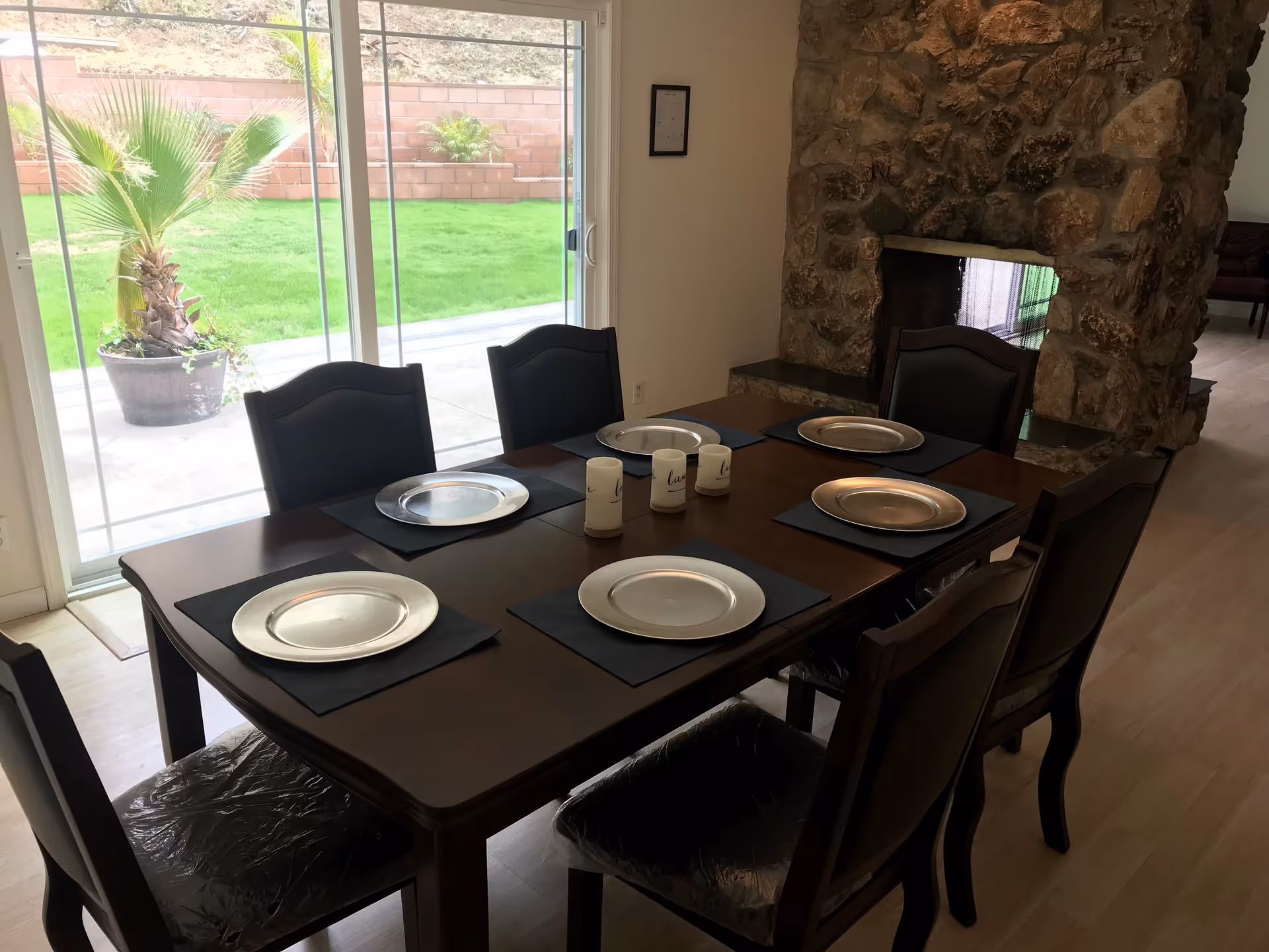 Dining room with a wooden table set for six with silver plates on black placemats, three candles in the center, a stone fireplace in the background, and a sliding glass door showing a green lawn and potted plant outside.