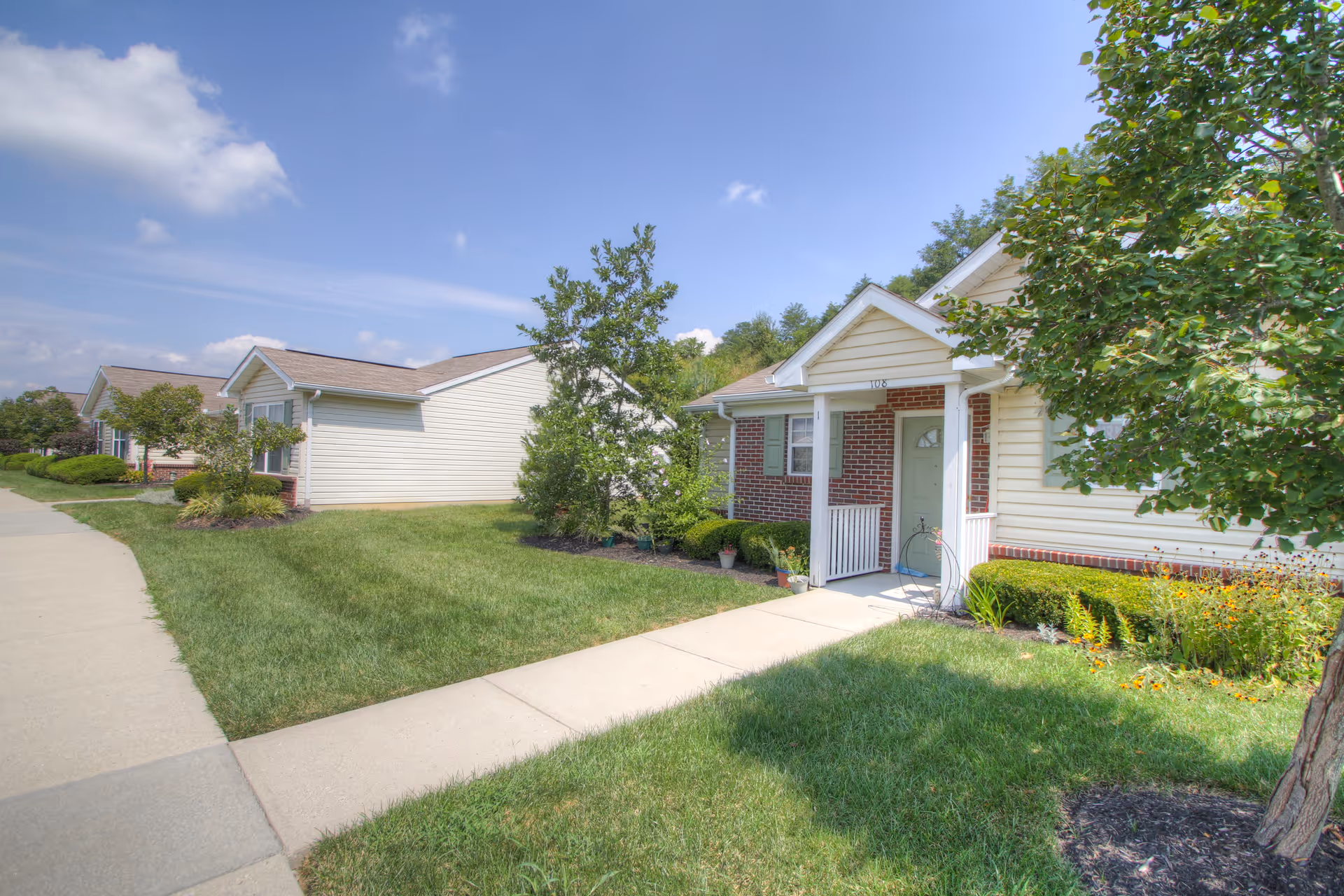 Exterior view of single-story residential buildings with beige siding and brick accents, surrounded by green lawns, trees, and shrubs under a partly cloudy blue sky.