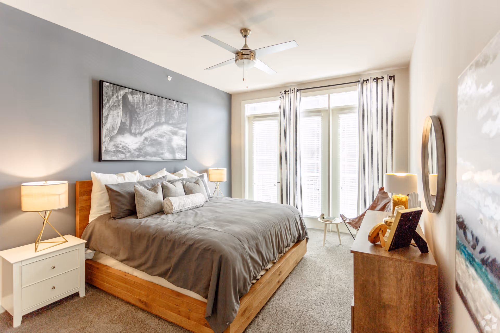 A bright and modern bedroom with a large wooden bed featuring gray bedding and multiple pillows. There are two bedside tables with lamps, a ceiling fan, a dresser with decorative items, a round mirror on the wall, and large windows with striped curtains letting in natural light.