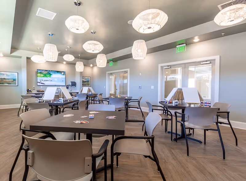A bright, modern activity room with multiple tables and chairs arranged for group activities. Each table has art supplies and small easels set up for painting. The room features hanging pendant lights, a large TV on the wall displaying a beach scene, and framed artwork. The flooring is wood-style, and there are glass double doors and exit signs visible.