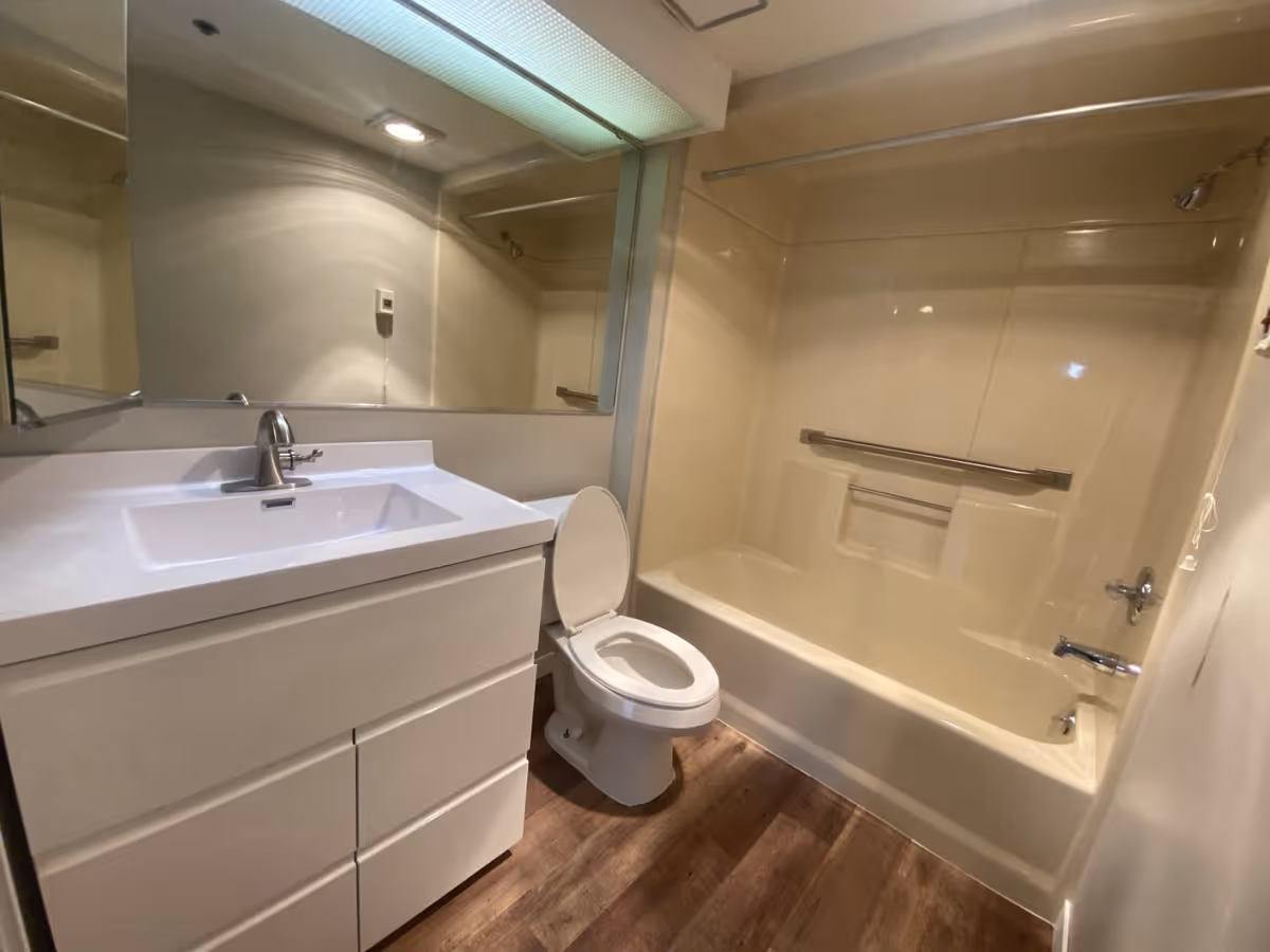 A bathroom with a white vanity sink, a toilet with the lid open, and a bathtub with a showerhead and grab bar. The floor has wood-like flooring and there is a large mirror above the sink.