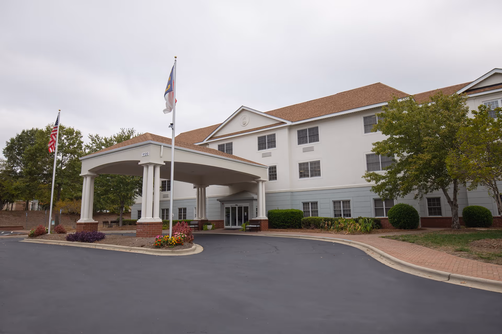 Exterior view of The Charlotte senior living facility showing a three-story building with a covered entrance, two flagpoles with flags, landscaped greenery, and a paved driveway.