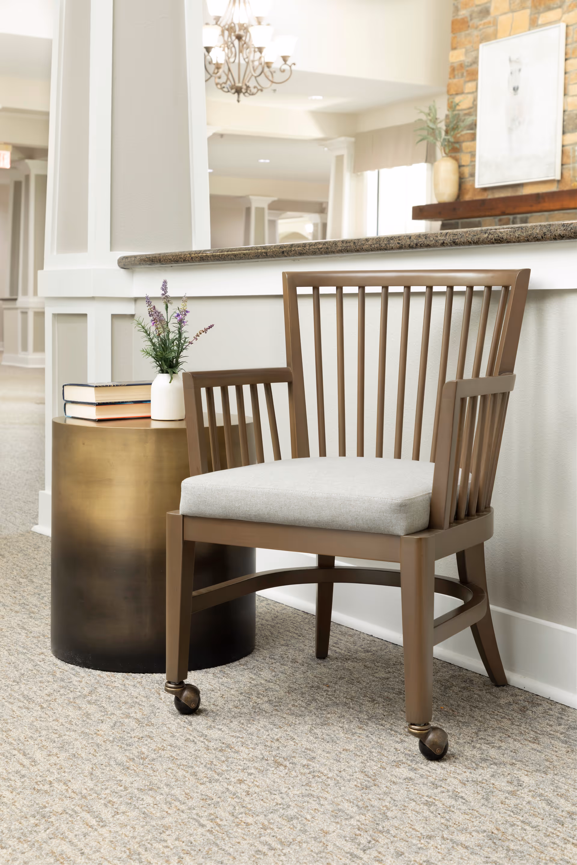 A wooden chair with a light gray cushion on wheels is positioned next to a round metallic side table holding a small white vase with purple flowers and two stacked books. The background shows a bright, well-lit interior space with a granite countertop, white walls, and a stone fireplace with a framed picture above it.