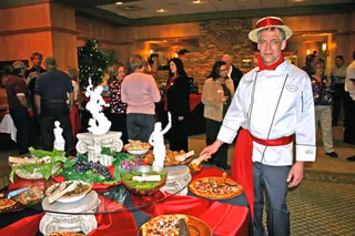 A man dressed as a chef stands next to a buffet table with various foods including pizza, grapes, and salad in a room where several people are socializing in the background.