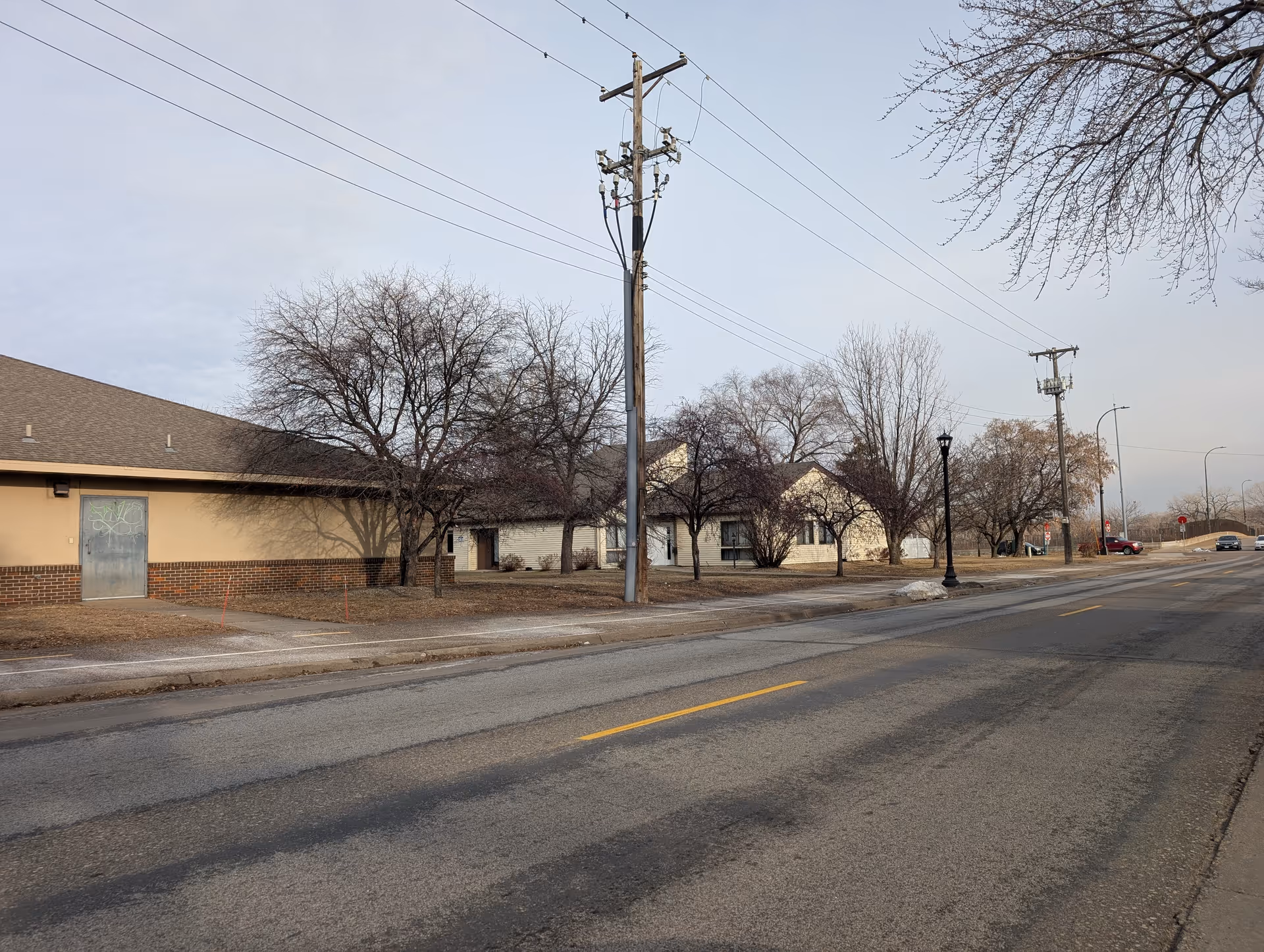 Street-front view of low-rise facility buildings with leafless trees, utility poles, and an empty road under a gray sky.