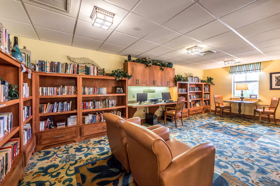 A cozy library room with wooden bookshelves filled with books, two leather armchairs, a desk with two computers, and a small table with two chairs near a window with a striped valance. The room has a patterned carpet and soft yellow walls with decorative plants on top of the shelves.