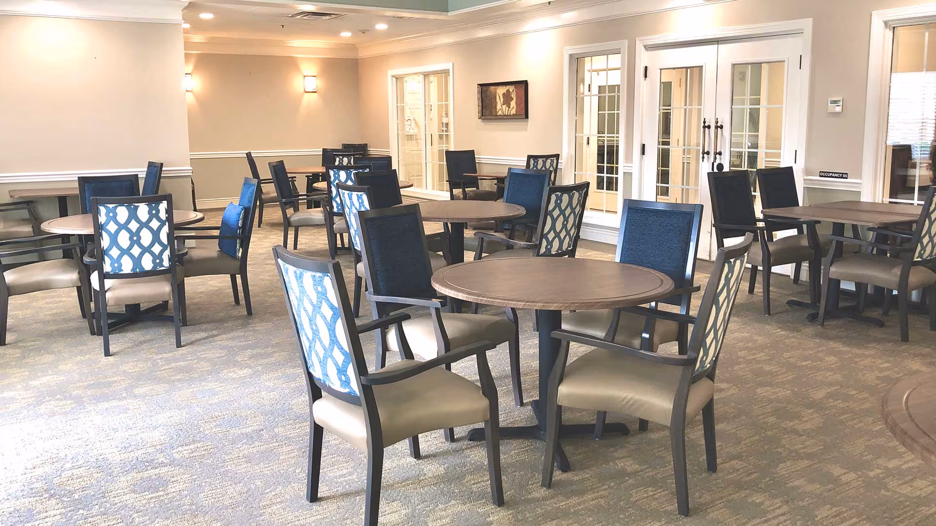 A spacious dining room with round tables and chairs featuring blue and beige upholstery, illuminated by soft lighting, showcasing a welcoming atmosphere.